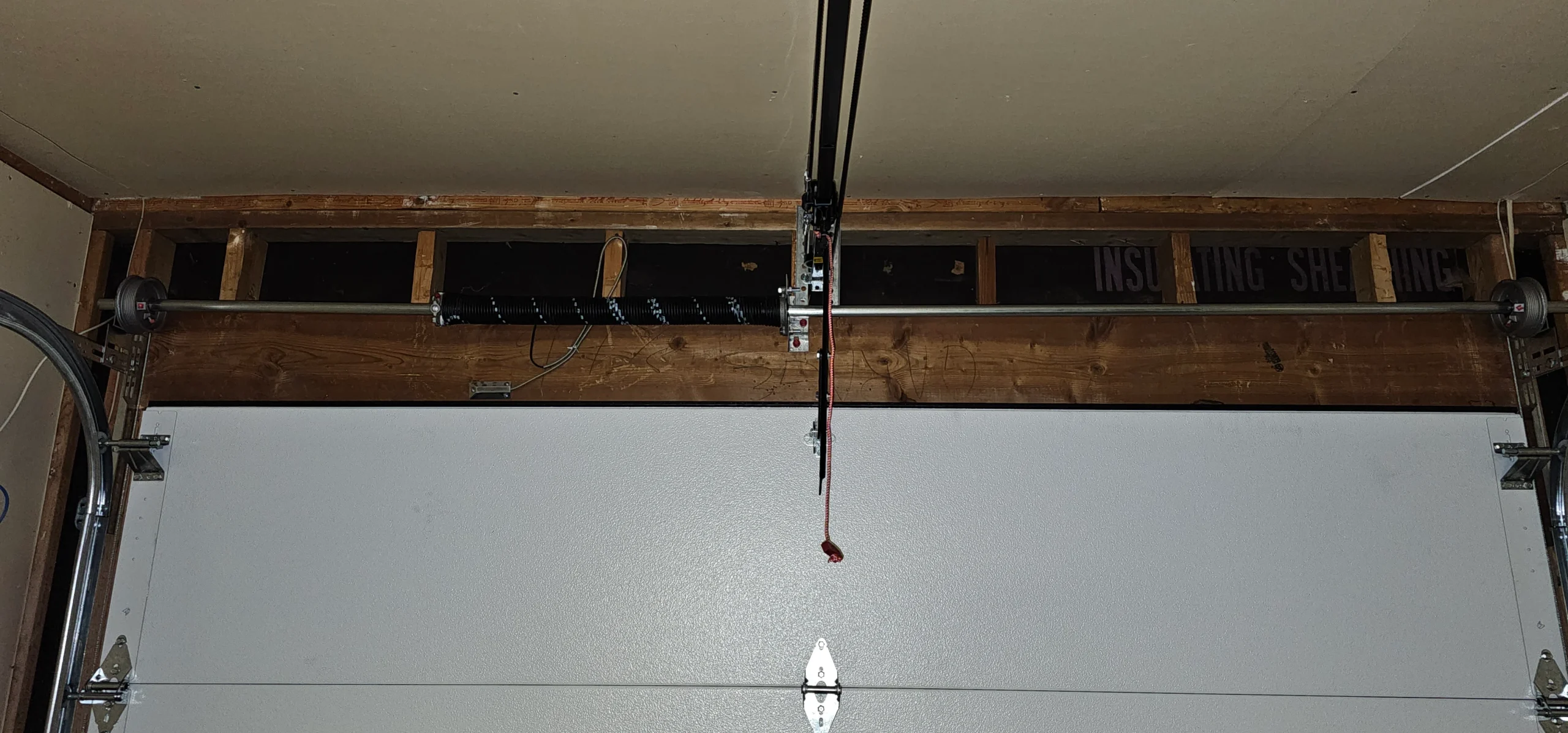 An overhead view of a garage door lifting system captures a single, heavy-duty torsion spring mounted horizontally on a steel shaft with cable drums at each end, cleanly demonstrating the mechanisms discussed in Role of Torsion Springs in Garage Doors
